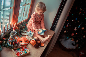 Cute little girl is dressing up a tiny Christmas tree and trying on New Year's animal masks. Image with selective focus and toning. 