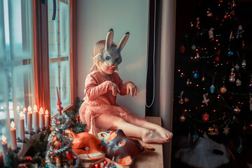 Cute little girl is dressing up a tiny Christmas tree and trying on New Year's animal masks. Image with selective focus and toning. 