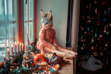 Cute little girl is dressing up a tiny Christmas tree and trying on New Year's animal masks. Image with selective focus and toning. 
