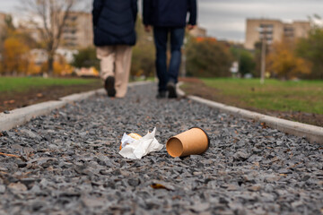Disposable coffee cup and a piece of croissant thrown away on the ground  in park with young couple walking in the background. Environmental pollution and litter concept.