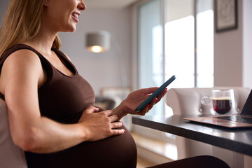 Pregnant Woman Working From Home On Laptop Looking At Phone Sitting At Table Touching Stomach