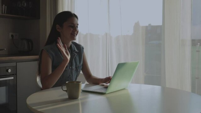 Image of young cheerful happy positive cute beautiful business woman sitting at home and using laptop computer talking on video call and waving hand while sitting at table in kitchen.