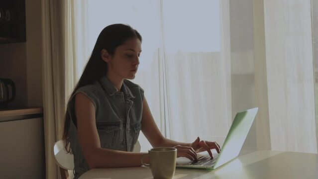 Image of young cheerful happy positive cute beautiful business woman sitting at home and using laptop computer talking on video call and waving hand while sitting at table in kitchen.