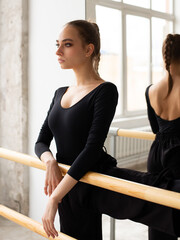 Modern female dancer in black outfit standing in front the mirror leaning on wooden handrail while practices in a dance studio - 541452465
