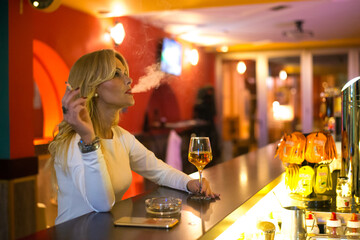 Middle aged blonde elegant woman sitting in the bar, drinking wine and smoking cigarette at night