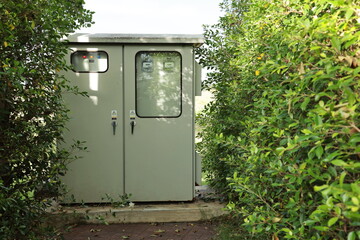 Outdoor electrical control cabinet mounted on a concrete base in the grass behind the green hedges.