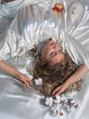 A beautiful young blonde in a stylish silk white dress and necklace poses in the studio lying on the bed. Top view.