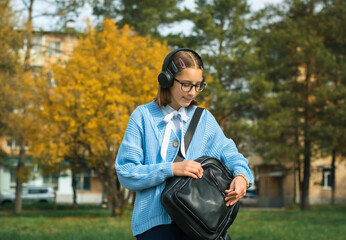 A young schoolgirl with a backpack in headphones is walking down the street listening to music on a sunny day.