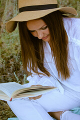 Kyiv Ukraine. August 1, 2021 Young cheerful woman, girl in white clothes and hat reading a book at a picnic. Recreation in nature in the park in sunny summer autumnal day. Female tourist in lotus pose
