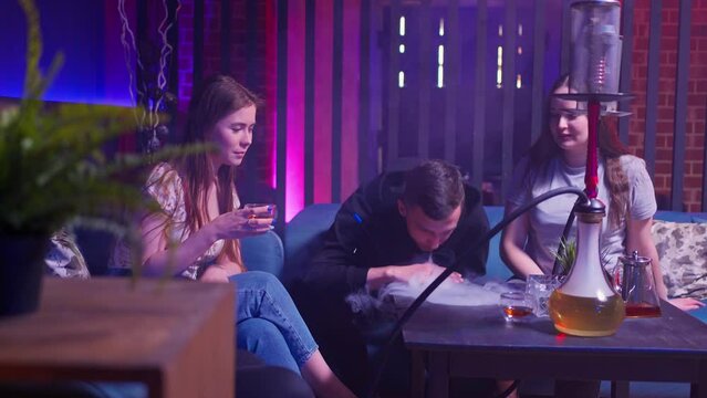 a company of young friends, a young guy and two girls are sitting on a sofa in a hookah room, smoking a hookah and talking. the guy blows hookah smoke on the table