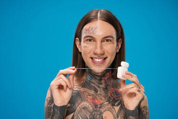 cheerful and tattooed man holding dental floss while looking at camera isolated on blue