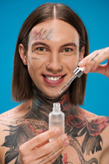 smiling and tattooed man holding bottle with moisturizing serum isolated on blue