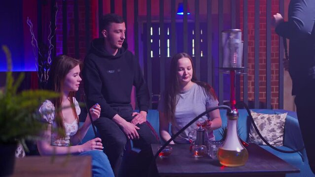 a company of young friends sit on a sofa in a hookah lounge, smoke a hookah and talk