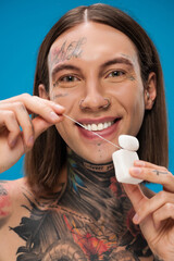 Obraz na płótnie Canvas cheerful and tattooed man holding dental floss isolated on blue