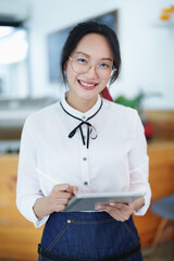 Starting and opening a small business, a young Asian woman showing a smiling face holding a tablet in an apron standing in front of a coffee shop bar counter. Business Owner, Restaurant, Cafe concept