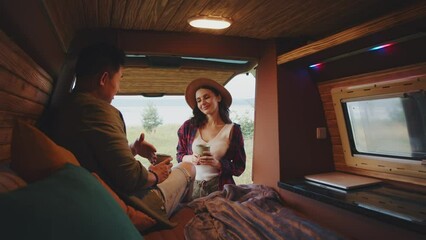 Cozy interior of modern comfortable camping custom design car. Bed with colorful pillows. Beautiful Caucasian woman holding cup of coffee and talking to boyfriend. Romantic. Tourism. Weekend. Daytime
