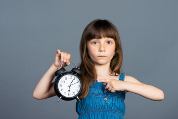 A little preschooler girl holds a round alarm clock in her hands and shows the time with her index finger. Isolated on a gray background. Time to study and don't be late for school