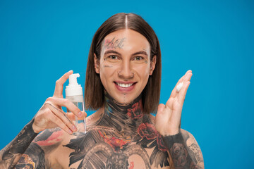 joyful and tattooed man holding bottle with cleansing foam isolated on blue