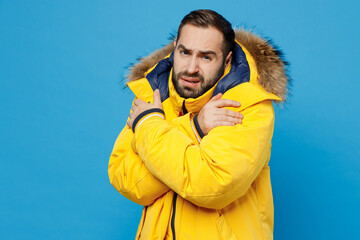 Young frozen sad upset man 20s wearing yellow puffer down jacket with fur hood hug himself trying...