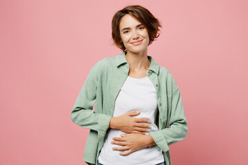 Young smiling cheerful fun happy woman 20s she wear green shirt white t-shirt holds hands on belly look camera isolated on plain pastel light pink background studio portrait. People lifestyle concept. - obrazy, fototapety, plakaty