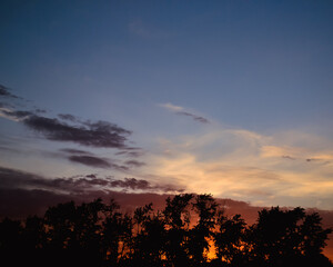 Morning glow illuminates the clouds on a beautiful colorful sky above the silhouettes of trees at dawn