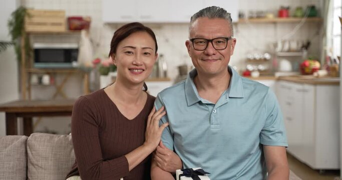 portrait of asian retired father and adult daughter looking at camera with Father’s Day gift in living room at home. the smiling woman hugs the man and leans on his shoulder