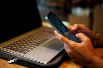 Close-up of woman's hand using smartphone while working on laptop in a coffee shop.