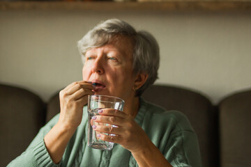 Caucasian senior woman in mint knitted sweater taking blue capsule in her mouth and drinking water from glass at home.