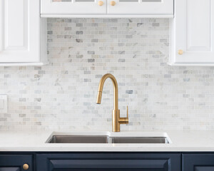 A kitchen sink detail shot in a white and blue kitchen with a gold faucet, marble countertop, and...