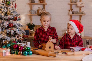 boy and girl in red christmas pajamas prepare a gingerbread house and decorate it with icing at home