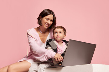 Cheerful young woman and little boy, mother and son using laptop isolated on pink studio background. Mother's Day celebration. Concept of family, childhood, motherhood