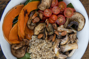 Top view of a nutritious salad with grilled vegetables. Vegetarian salad with grilled pumpkin, mushrooms, spinach, barley, almonds and cherry tomatoes, in a white bowl on the restaurant wooden table.