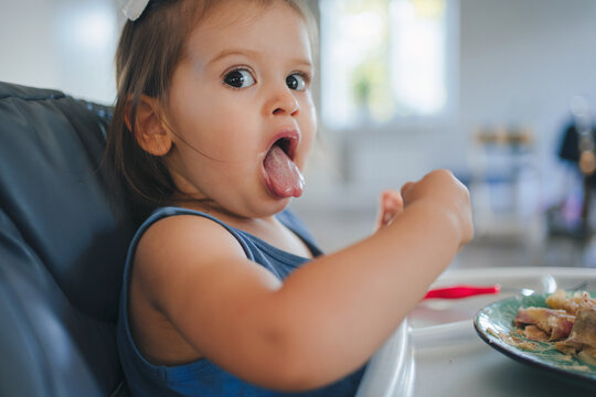 Baby girl sitting with mouth open and tongue sticking out while eating with her hands from the plate full of tasty food. Healthy eating for kids children