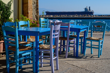 Selective focus on the close blue wooden table and chair at typical fish tavern at fishing port. Big ship on background. Aegean island Chios in Greece on an autumn day. Greek holidays and destinations