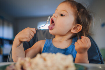 Baby girl sitting at the children's table, eating with appetite and licking the spoon. Healthy...