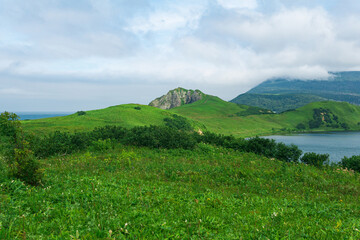 Obraz premium natural landscape of Kunashir island with grassy hills, volcanic rocks, volcano in the clouds, lagoon and ocean in the distance