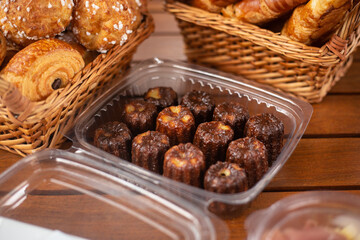 A transparent plastic box full of french traditional canelés dessert next to two wooden baskets...