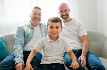 Happy two man couple with adopted child sit on sofa at home