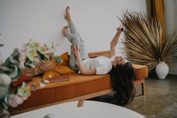 Young hispanic brunette woman laying on orange sofa with legs raised on the wall, speaking with...