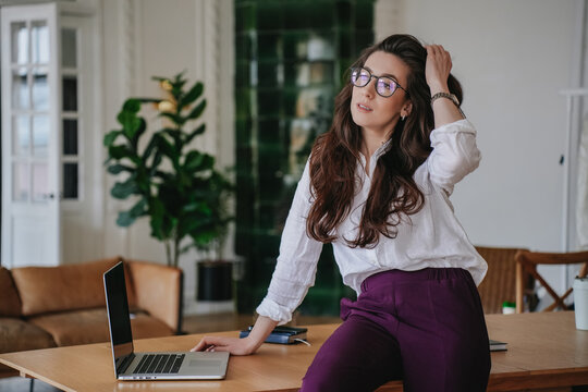 Empowered Hispanic Brunette Woman In Glasses With Wavy Loose Hair In White Shirt Looking Aside Confidently Over Blurry Office. Portrait Of Smart Student At Home. Business And Education