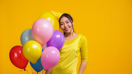 Fototapeta na wymiar happy asian woman in party cap holding colorful balloons isolated on yellow