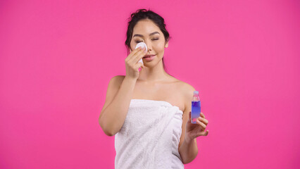 young asian woman in towel holding cotton pad with micellar water while cleansing face isolated on pink