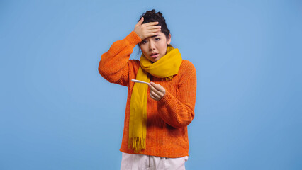diseased asian woman in sweater and scarf holding digital thermometer and touching forehead...