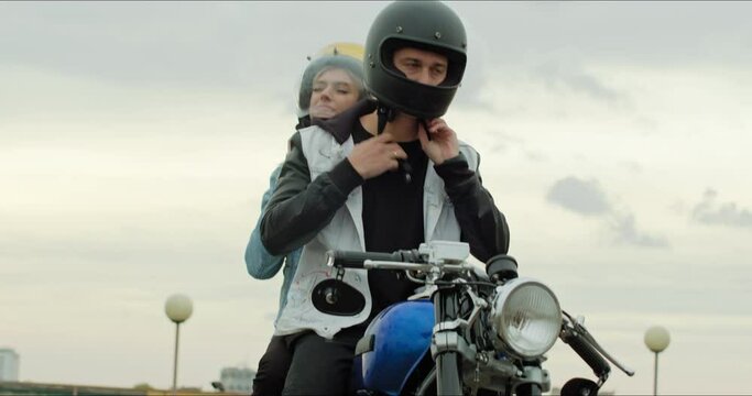A young stylish couple of bikers sit on a bike standing in a public city parking. The guy and the girl put on motorcycle helmets. Love and safety