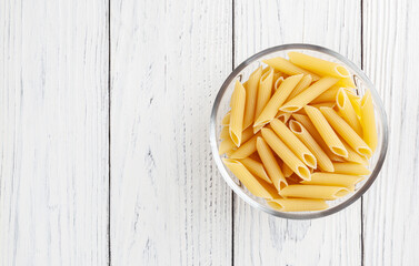 Uncooked penne pasta in glass bowl on white wooden background