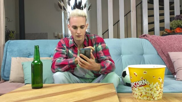 Nervous young man looking at smartphone, rejoicing score of soccer match, having profit bet on bookmaker online app, pointing on green chroma key screen, sitting on sofa indoors. Gambling concept