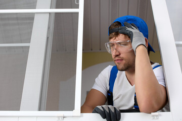 An upset window installer thinks with closed eyes how to solve the problem by holding his head with his hand. Leaning his head on his hand, a professional master reflects on the situation