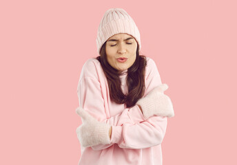 Beautiful brunette woman in warm pastel pink winter hat, sweatshirt and mittens standing on light pink background, feeling very cold, freezing, hugging herself and shivering with funny face expression
