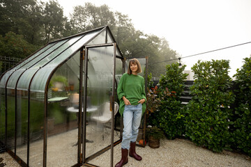 Portrait of young woman stands near greenhouse for growing plants at backyard. Gardening, leisure time in garden concept