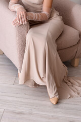 Beautiful fashionable young woman in elegant beige dress and gloves sitting in armchair at home